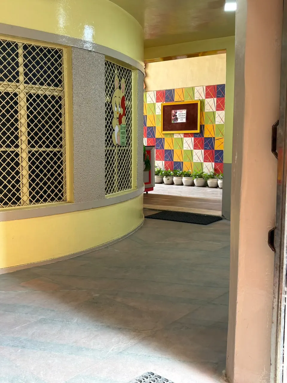 Yellow curved entrance corridor leading to a colourful tiled wall and potted plants — Children Glory High School, Morar, Gwalior
